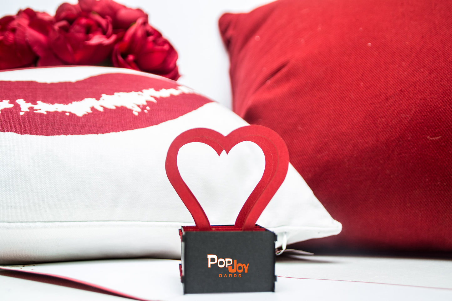 Pop-up card featuring three red heart-shaped arcs popping out of a black box in the middle, with the card set in front of white and red pillows