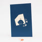 Dark blue card with silhouette of baby peeking out of window and two diaper pins on front cover