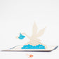 Pop-up card with white crane flying through blue and white clouds and carrying baby in blue polka dot cloth bundle