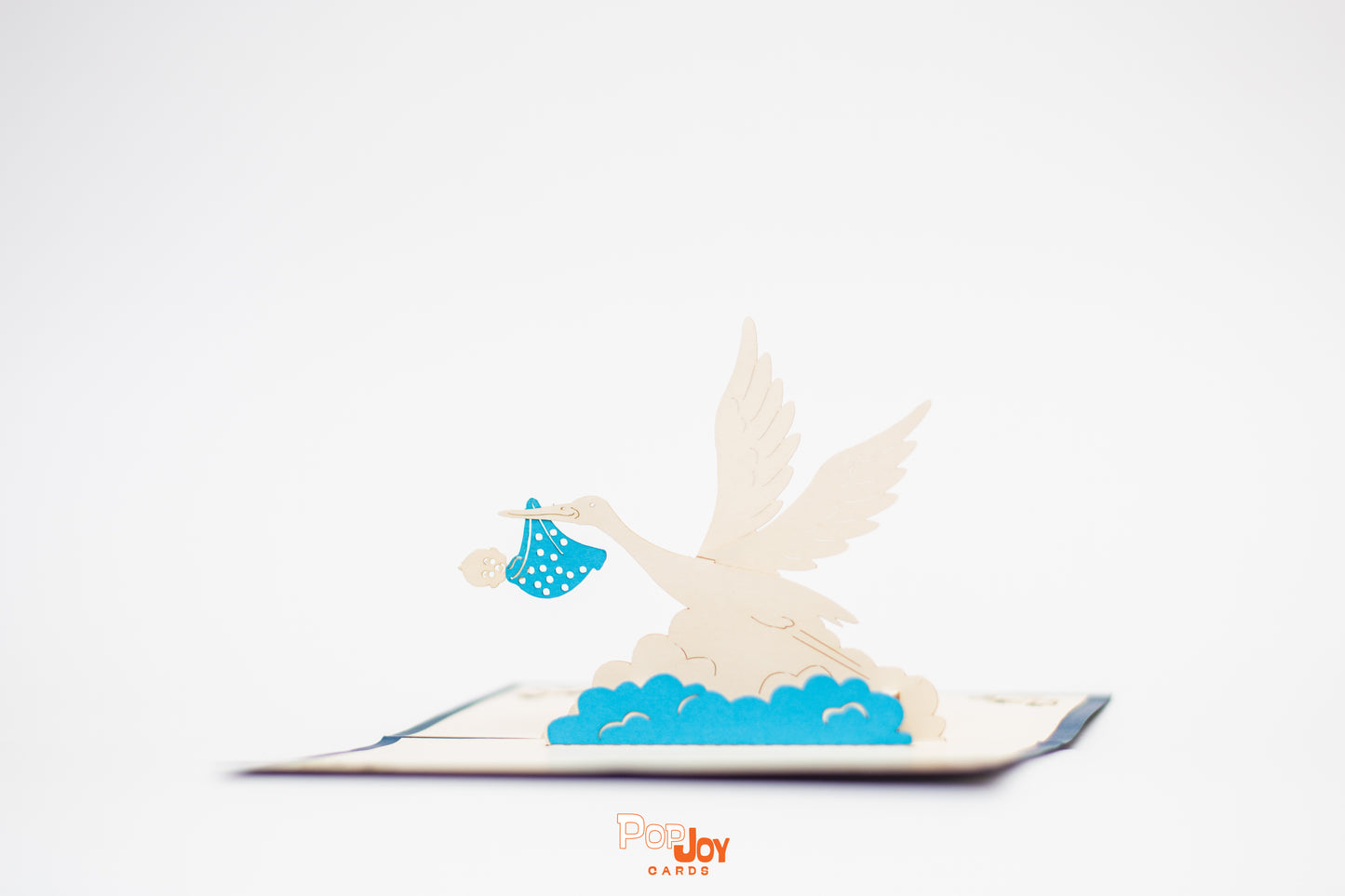 Pop-up card with white crane flying through blue and white clouds and carrying baby in blue polka dot cloth bundle