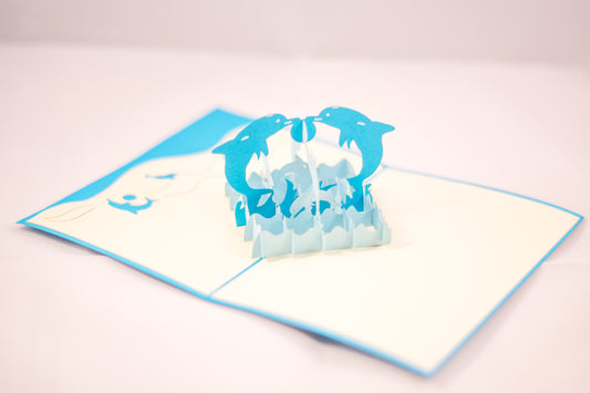 Pop-up card showing two blue dolphins playing with a ball in the middle, while surrounded by waves and other dolphins