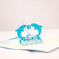 Pop-up card showing two blue dolphins playing with a ball in the middle, while surrounded by waves and other dolphins