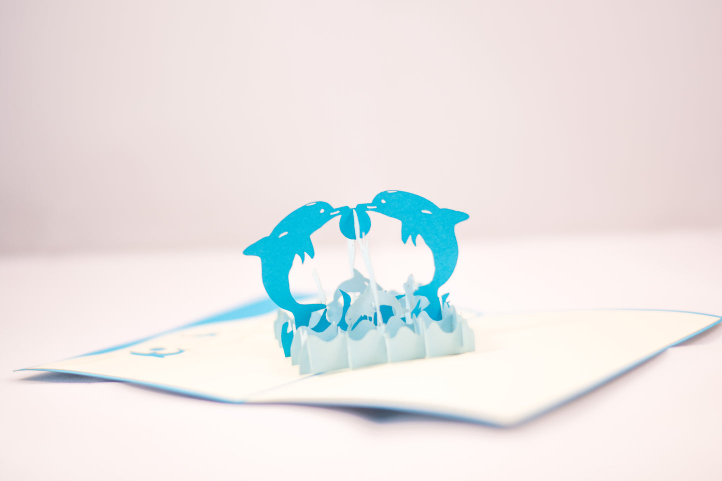 Pop-up card showing two blue dolphins playing with a ball in the middle, while surrounded by waves and other dolphins